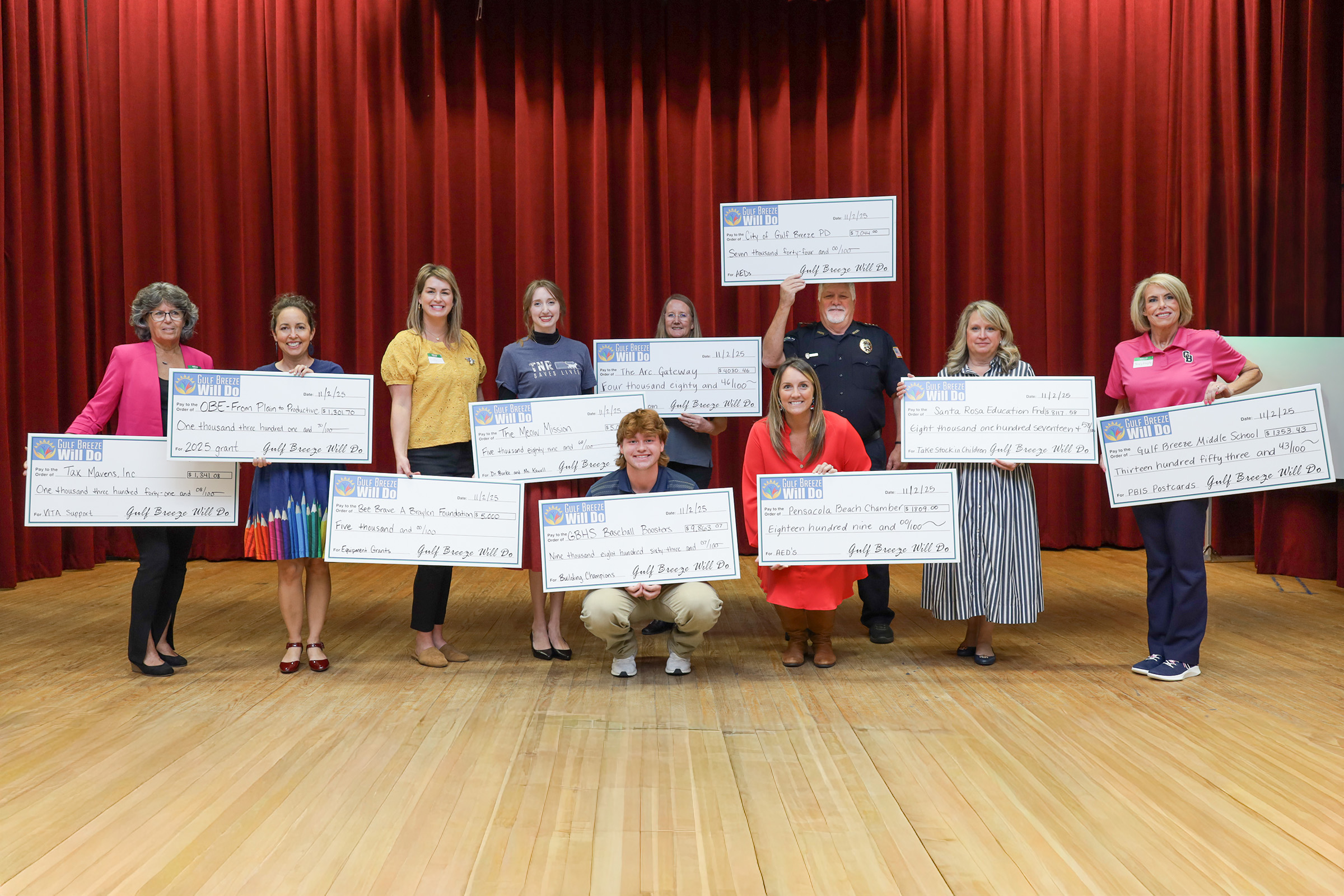 A group of people standing in front of a red curtain and all holding oversized checks from Gulf Breeze Will Do