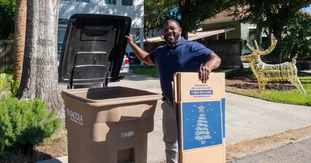 A man holding a box with holiday graphics on the side, about to put the box in a recycling bin from the City of Pensacola.