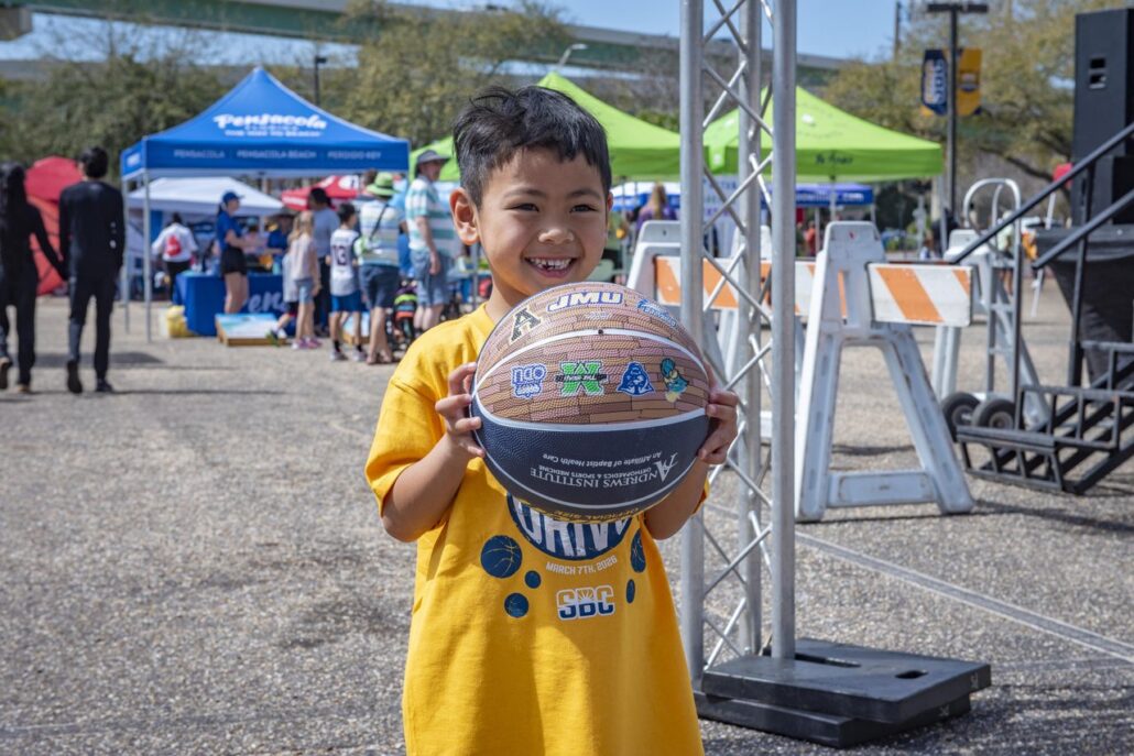 Child in a yellow shirt holding a basketball at the free fasn fest at the 2026 Sun Belt Basketball Championships in Pensacola, Florida.