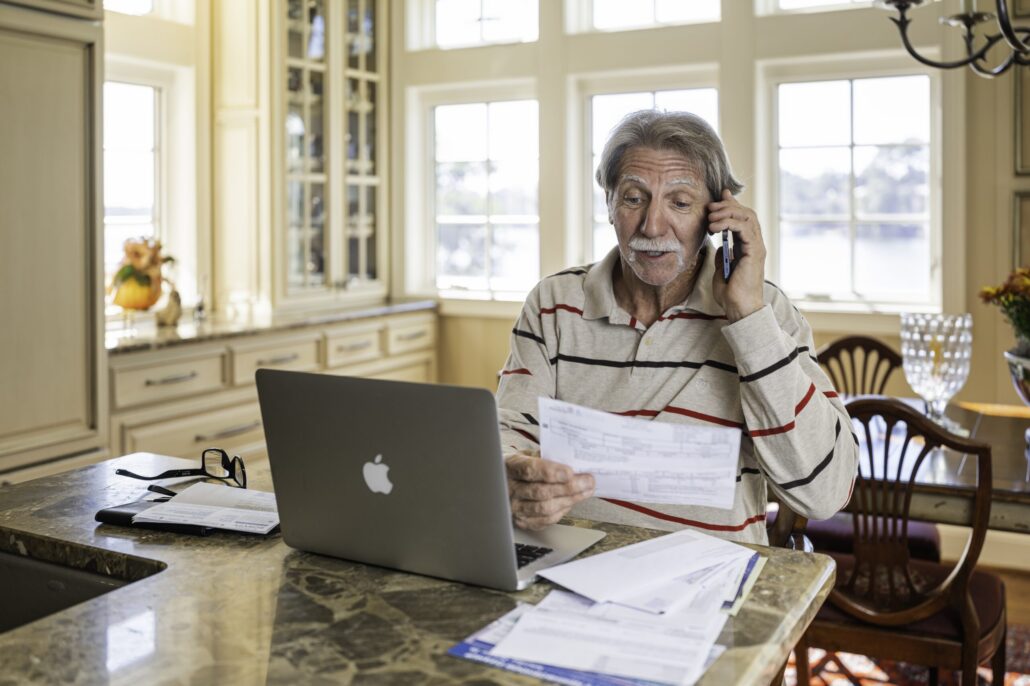 Man with gray hari sitting at a kitchen table with a laptop and papers. He is holding a phone up to his ear.
