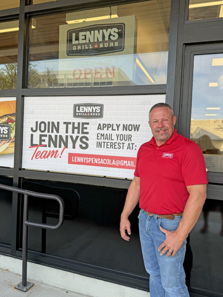 Man in a red shirt stands in front of the store windows for Lennys Sub shop in downtown Pensacola.